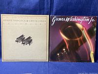Front view of both record sleeves side-by-side on blue background. Left sleeve is beige with piano graphic, titled 'An Evening With Herbie Hancock & Chick Corea In Concert'. Right sleeve shows Grover Washington Jr. with a saxophone image on a purple background.