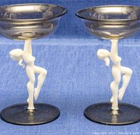 Pair of clear smoky glass cocktail glasses with frosted white nude lady figure stems, side view on blue fabric background.