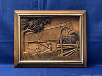 Frontal view of wooden bas-relief plaque showing detailed rural scene with covered bridge, tree, fence, and fishing children in warm wood tones inside a layered wood frame.