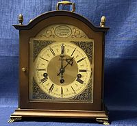 Front view of vintage Tempus Fugit Forestville German mantel clock showing clock face with Roman numerals, decorative metal corners, brass handle on arched wooden top, and brass feet.