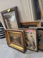Framed artwork group showing multiple pieces leaning against a wall, various sizes and styles visible, frames show age and wear.