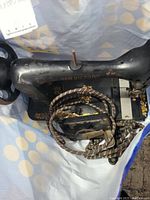 Black New Victory sewing machine with gold floral decorations, partially visible hand crank, and coiled fabric-covered foot pedal cable on top of machine base.