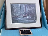 Photo of framed print depicting moose crossing a river amid a forest scene, showing the full print within a wood frame with visible damage.