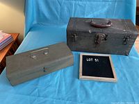Two vintage metal toolboxes side by side on a blue cloth background; one black with a brown leather handle and latch, the other long and olive green with a horizontal handle.