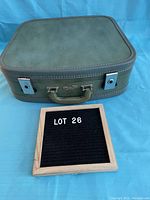 Photo showing the closed green hardcase suitcase with brown handle and silver latches, placed on a blue background with lot label beside it