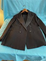 Black double-breasted tuxedo jacket with peak lapels laid flat on blue cloth with hanger inside, showing front button closure and pockets.