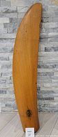 Full view of the wooden propeller blade standing upright against a stone-patterned wall.