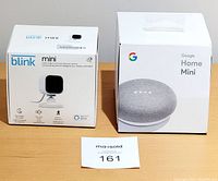 Boxes of Blink Mini camera and Google Home Mini speaker side by side on a wooden surface with MaxSold lot number tag.