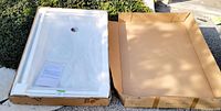 Shower tray packaged in cardboard box, white acrylic surface visible with drain hole, protective wrap covering tray.