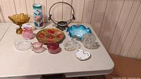 Table with assortment of antique glass items including colorful vases, bowls, clear glass dishes, and metal handled basket.