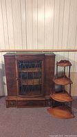 Antique wooden hutch with glass door and two drawers next to a 4-tier corner shelf in a room with wood panel walls.