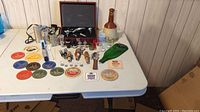 Wide view of the entire bar accessories collection on a white folding table, showing bottles, stoppers, coasters, and other items.