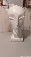 Front view of off-white stylized head-shaped ceramic vase/planter showing facial features and square base.