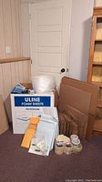 Photo showing a large white Uline box filled with thick polyethylene foam sheets, next to assorted mailing bags, rolls of tape, bubble wrap, and stacked cardboard boxes.