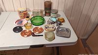 Wide view of the collection showing all items including ashtrays, tobacco tins, lighters and pipe on white table with wood panel background.