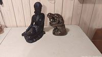 Photo of two sculptures on a white table in front of wood panel wall: a black matte seated woman figurine and a metallic finish thinking man figurine.
