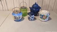 All five vintage biscuit barrels and ginger jars displayed together on wood surface, showing variety of shapes and colors.