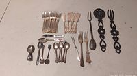 Overview of all cutlery items arranged on a table: multiple silver plated forks and spoons, hand-forged spoon and fork, ceramic-handled knives, decorative black metal utensils, Rolex and Royal Crown Derby spoons.