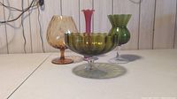 Four vintage glass pieces: green pedestal bowl, amber stemmed glass, pink bud vase, and green frosted vase.