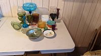 Wide shot showing assortment of colorful vintage and antique glassware on white table, including bowls, glasses, and a decorative plate.