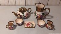 Photo of all seven Wade Harvest Ware pieces including teapot, creamer, sugar bowl, two small pitchers, and small rectangular dish with copper luster and floral motifs.