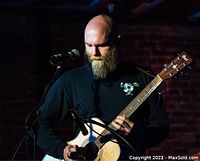 Photo shows Basil Park playing acoustic guitar on stage with microphone and brick wall background.