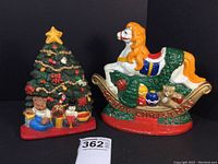Pair of vintage cast iron door stops: Christmas tree and rocking horse with 'Merry Christmas' base, shown on black background with lot number.