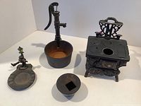 Four cast iron miniature items displayed on white surface: the stove, water pump with bucket, ash receiver with figurine, and small bowl with lid.