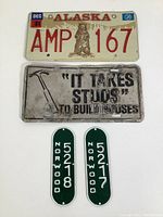 Top-down photo showing all three items: Alaska vintage license plate with bear, humorous metal license plate with hammer, and two Norwood green oval tags.