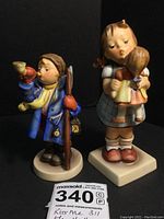 Two Goebel Hummel figurines side by side on black background with lot tag. Girl figurine is taller and holds a doll; boy figurine holds a horn and lantern.