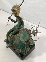 Side angle view of brass fishing boy sculpture seated on rock with wire fishing pole extending outward, showing patina and base.