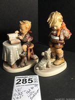 Two Hummel figurines on black background showing full front and side views. Little Bookkeeper seated at table with dog, Not For You standing with dog.