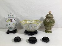 Two hand painted ginger jars, one white with colorful decorations and one green with raised floral motifs, one lotus bowl with gold floral patterns on a wooden stand, and five black wooden stands in front