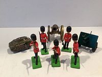 All items displayed together: five red-coated British soldier figures, two miniature cannons (brass and green painted), and green wind-up US Army tank toy.