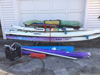 Front view of 3 stacked Mistral windsurfing boards with several sails and mast accessories in front on the ground.