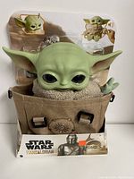 Front view of The Child plush toy in carrying satchel in original packaging showing character details and branding.