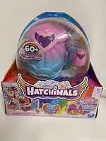 Front and angled view of Hatchimals CollEGGtibles Rainbow-cation Family Hatchy Home playset in original packaging showing main blue and pink egg-shaped container with purple heart design and included pink egg character