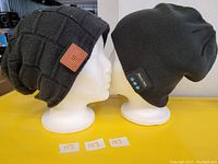 Two black Bluetooth-enabled toques displayed on white mannequin heads. The toque on the left has a checkered knit pattern and a brown leather patch. The toque on the right has a smooth knit with a small black control panel featuring blue buttons. Both appear new and are designed for wireless audio with charging capabilities.