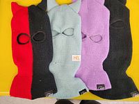 Four knit ski masks with horns laid out flat on a yellow surface; colors include red/black, light gray, purple, and black.
