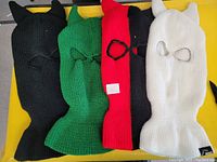 Photo of four knit ski masks in black, green, red, and white, each featuring horn-like protrusions and eye openings, laid flat on a yellow surface.