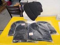 Four black knit toques individually packaged in plastic bags with one opened shown on mannequin head, displayed on a yellow table.