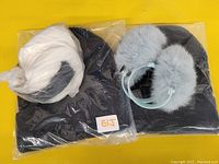 Two black knit toques sealed in plastic bags alongside two pairs of fuzzy earmuffs, one white and one light blue.