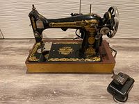 Side view of the black and gold Singer sewing machine showing intricate floral and Phoenix decal designs, foot pedal on the floor, and hand wheel detail.