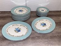 Overall view showing stacks of dinner plates, smaller plates, and two oval platters with white base and blue rose floral decoration around a light blue rim.