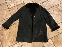 Front view of a black shearling coat laid flat on a tile floor, showing button closure, lapel collar, and visible shearling on cuffs and inside.