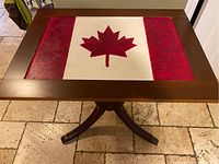 Top view of the rectangular wooden table showing hand-painted Canadian flag with red maple leaf and side red bands framed by brown wood, placed on tiled floor.