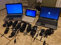 Three HP laptops with various power bricks laid out on a tile floor. Devices are powered on to show boot screens.