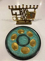 Two angled views showing the brass menorah with nine candle holders and the front side of the round brass Seder plate with six labeled oval brass sections and a central figure scene, turquoise patina base with grapevine details.