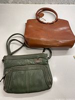 Two leather handbags, camel Rolfe with rounded handles on top and khaki green Tignanello with front zipper pocket and adjustable strap below, displayed side by side on white surface.