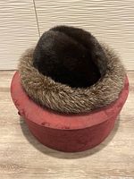 Dark brown mink fur hat with lighter fur trim inside red round hatbox on wooden floor, front view.